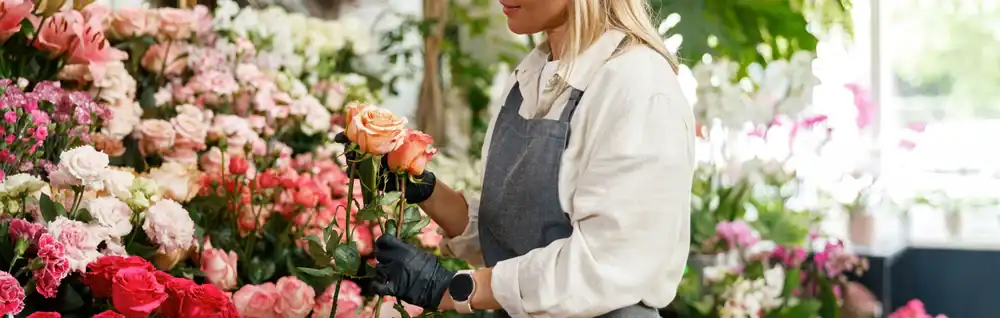 A florist wearing a gray apron and black gloves arranges pink and orange roses in a flower shop filled with various colorful blooms.