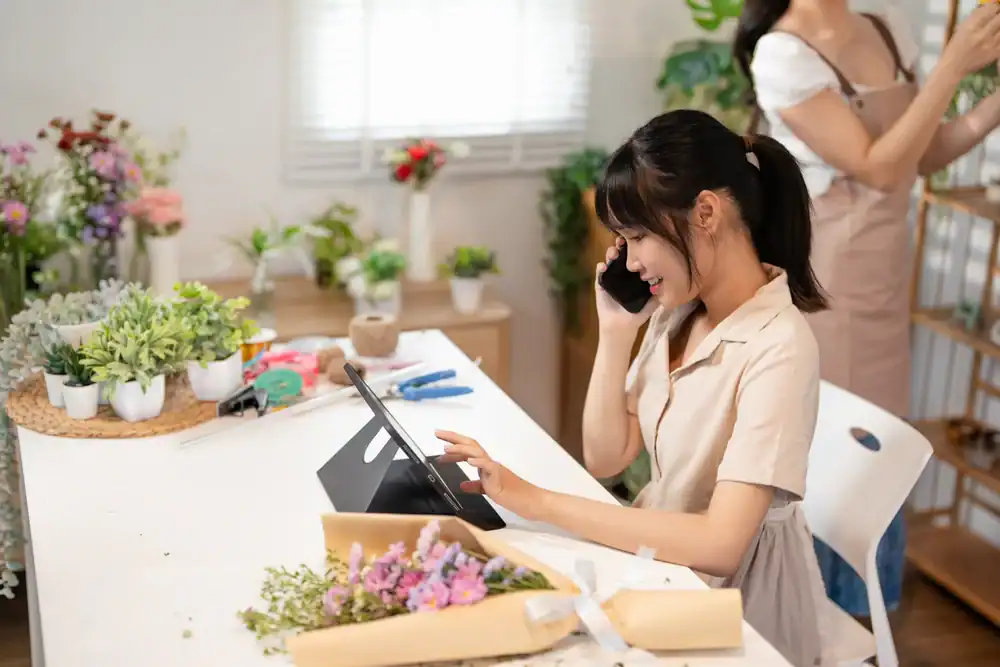 A woman sits at a table with flowers, talking on the phone and using a tablet, while another woman arranges plants in the background of a bright, cozy room filled with potted plants and floral supplies.