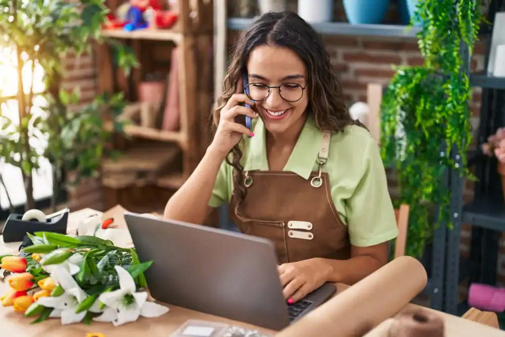 A woman wearing glasses and an apron sits at a table with a laptop, talking on her phone. She is surrounded by flowers and wrapping paper in a cozy, plant-filled workspace.