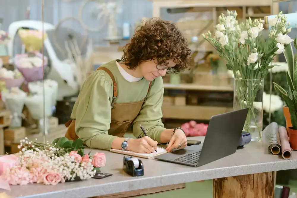 A florist with curly hair and glasses, wearing an apron, writes in a notebook while working at a laptop on a counter surrounded by flowers and floral supplies in a flower shop.