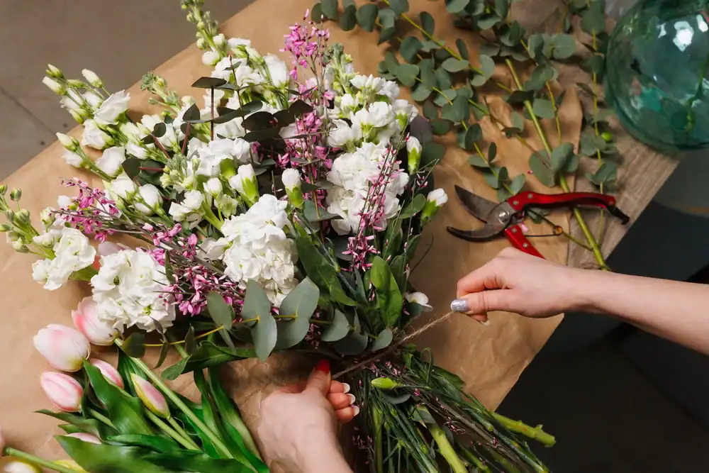 A person tying a bouquet of white, pink, and purple flowers with greenery on a table, next to tulips, pruning shears, and scattered eucalyptus leaves.