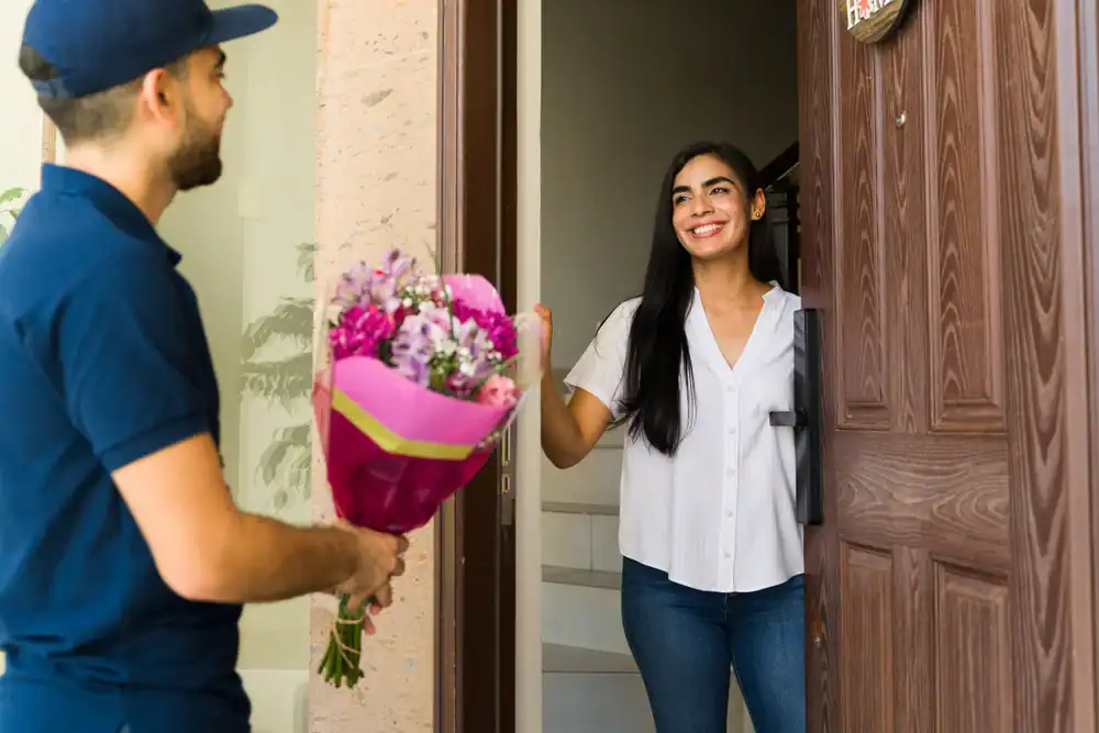 A smiling woman opens her front door to receive a bouquet of flowers from a delivery person wearing a blue uniform and cap.