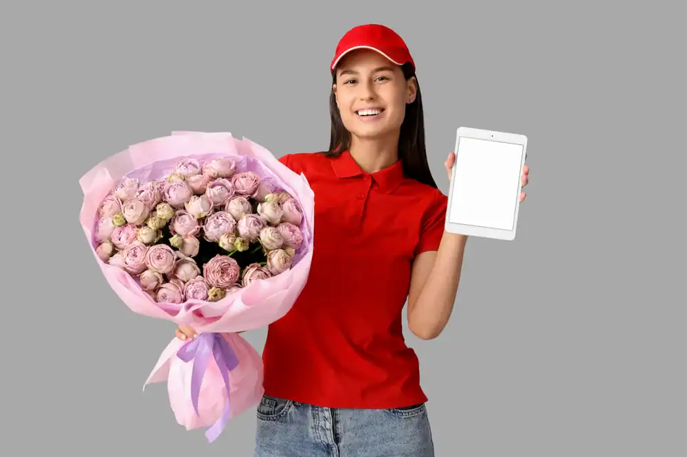 A smiling woman in a red cap and shirt holds a large bouquet of pink flowers in one hand and a blank tablet in the other, standing against a plain gray background.