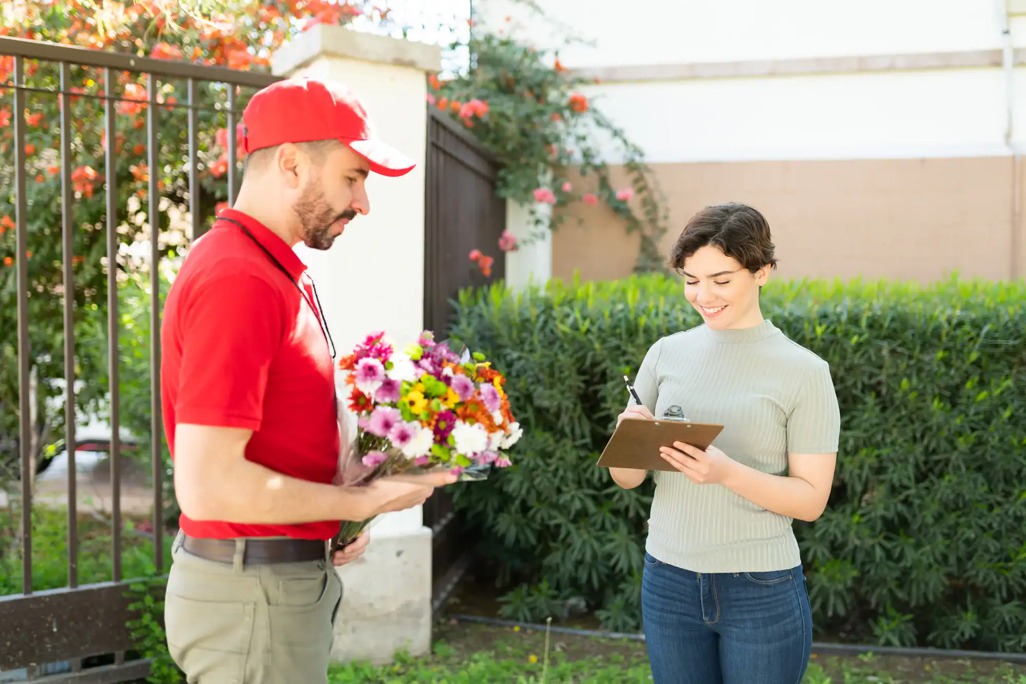 A florist in a red uniform and cap delivers a bouquet of colorful flowers to a smiling woman outdoors. The woman signs a clipboard while standing by greenery and a black gate.