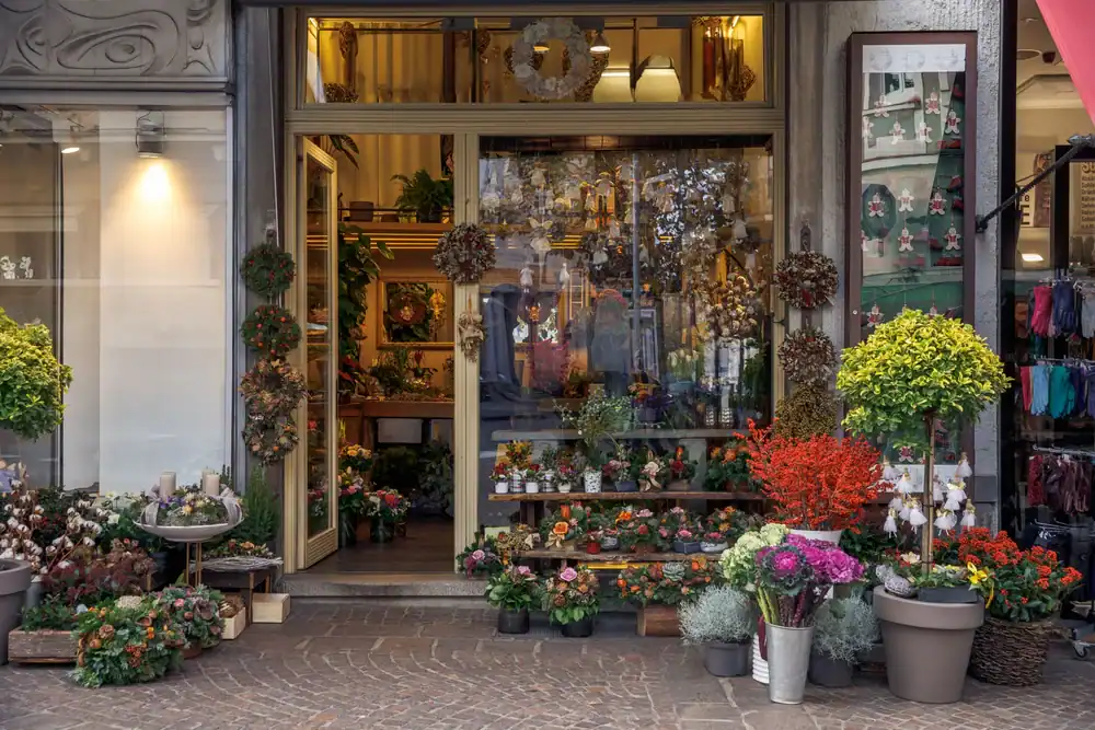 The entrance of a flower shop with potted plants, colorful flowers, and wreaths arranged outside and inside. The shop has large glass doors and windows, displaying more plants and floral decorations inside.