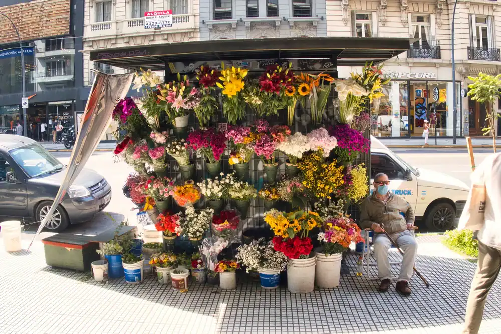 A colorful flower stand on a city sidewalk displays a variety of vibrant flowers in buckets. A man wearing a mask sits beside the stand, and urban buildings and cars are visible in the background.