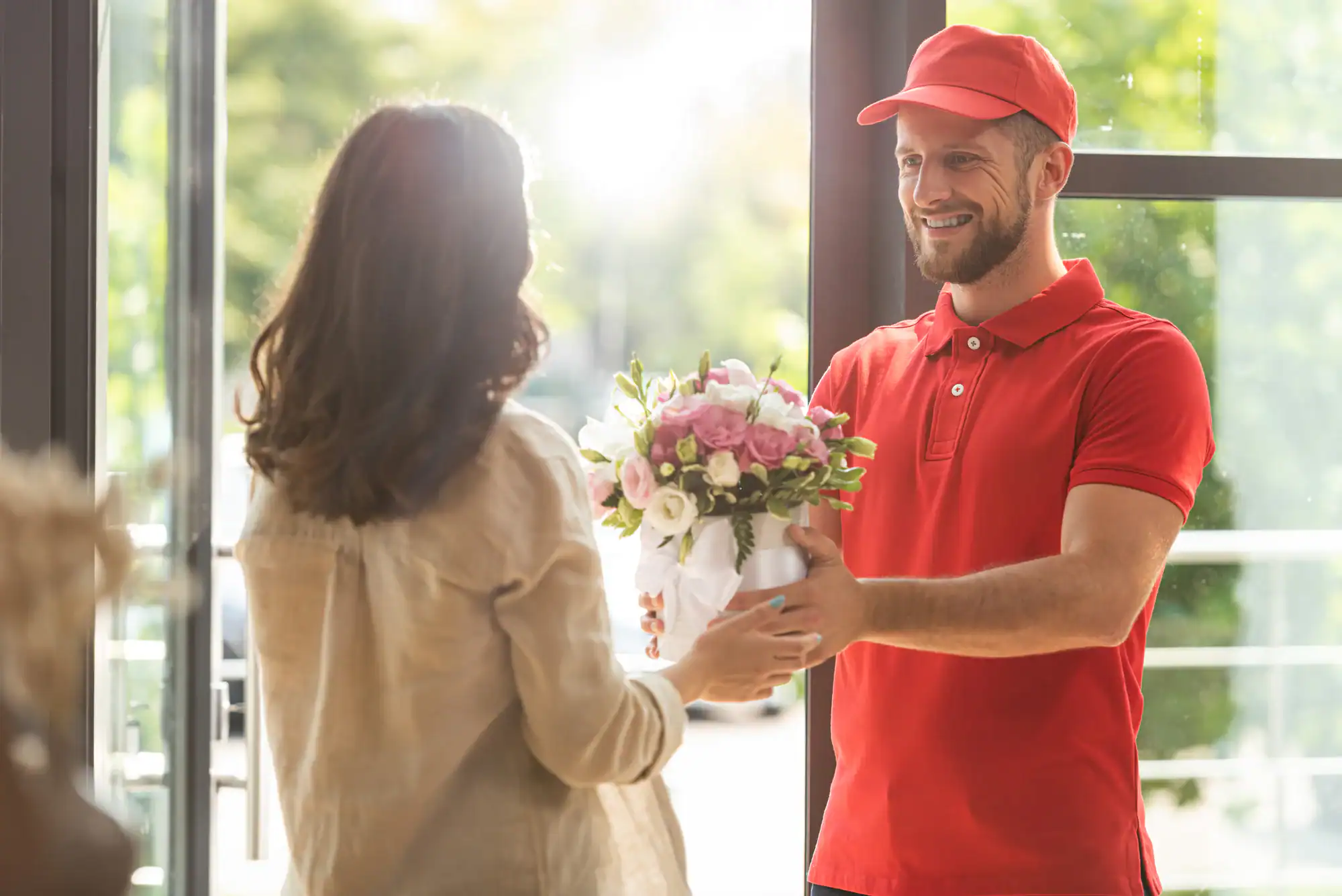 A smiling delivery person in a red uniform hands a bouquet of flowers to a woman at her door, with sunlight shining through the open doorway.