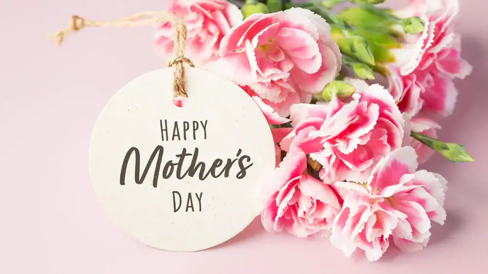 A round tag with "Happy Mother's Day" written on it is attached to a bouquet of pink and white carnations, set against a light pink background.