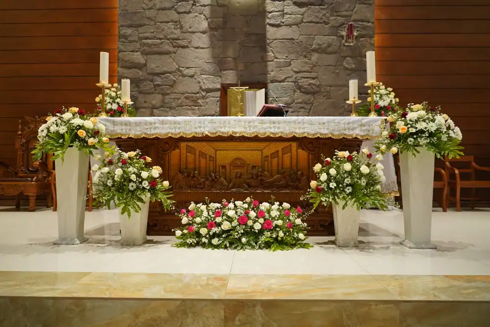 An ornate church altar decorated with white lace cloth, floral arrangements of white, pink, and yellow flowers in vases, and four white candles. The altar front features a carved scene; stone wall background.