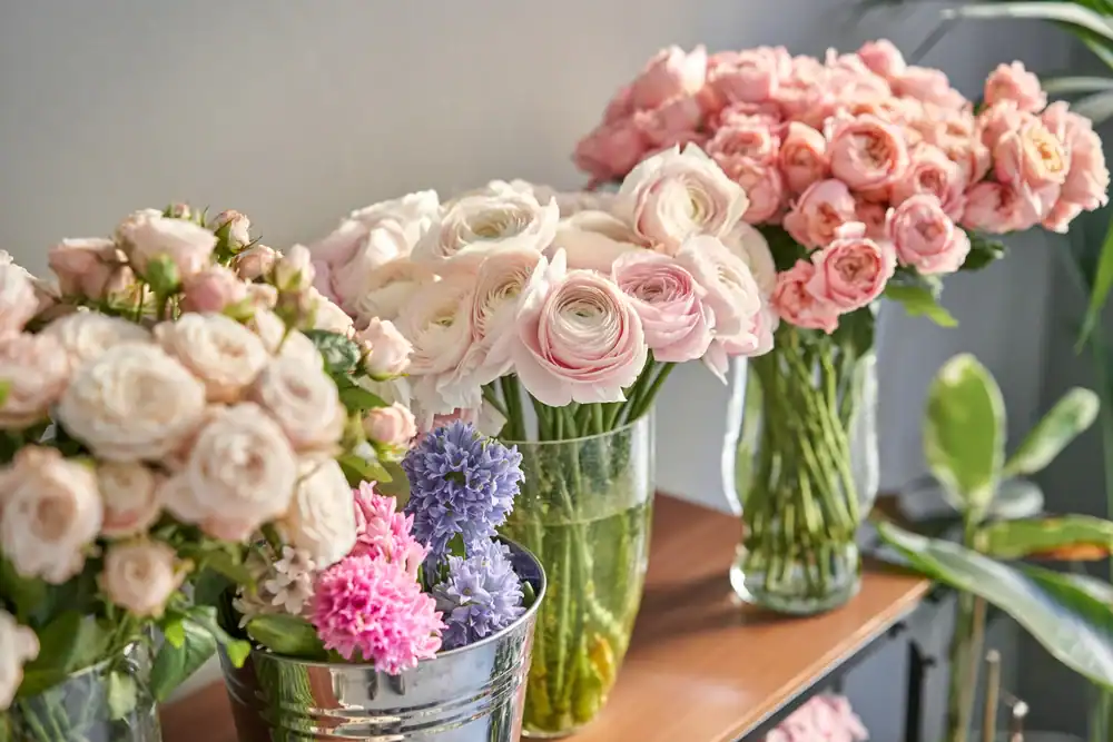 Several glass vases and a metal bucket hold bouquets of light pink and white ranunculus, roses, and purple hyacinths, displayed on a wooden shelf near a sunlit wall with green plants in the background.