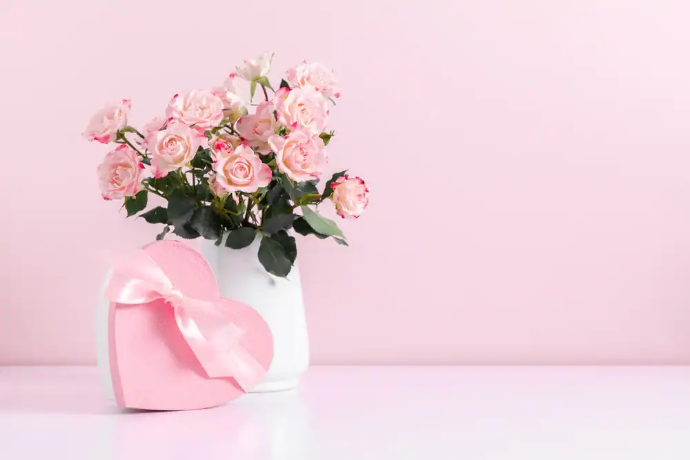 A white vase filled with pink roses sits next to a pink, heart-shaped gift box with a ribbon, all on a white surface against a pale pink background.