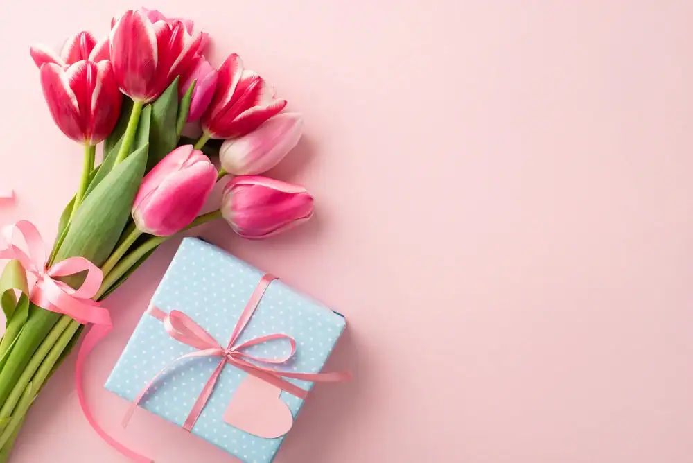 A bouquet of pink tulips and a blue polka dot gift box with a pink ribbon and heart-shaped tag are arranged on a pink background.