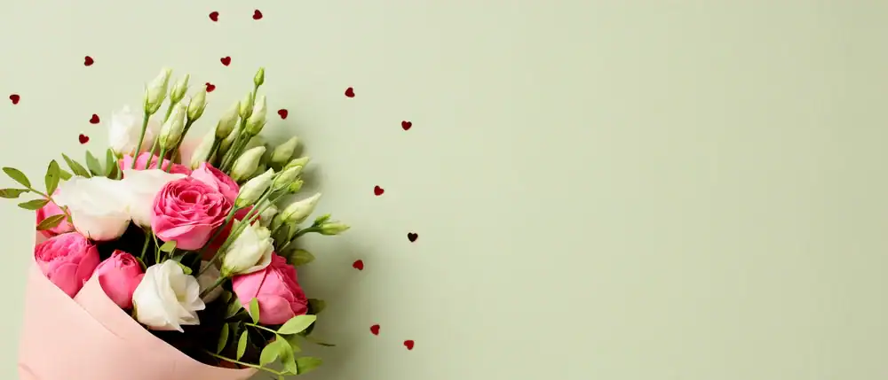 A bouquet of pink and white roses and lisianthus wrapped in pink paper is on the left side, with small red heart-shaped confetti scattered on a light green background.