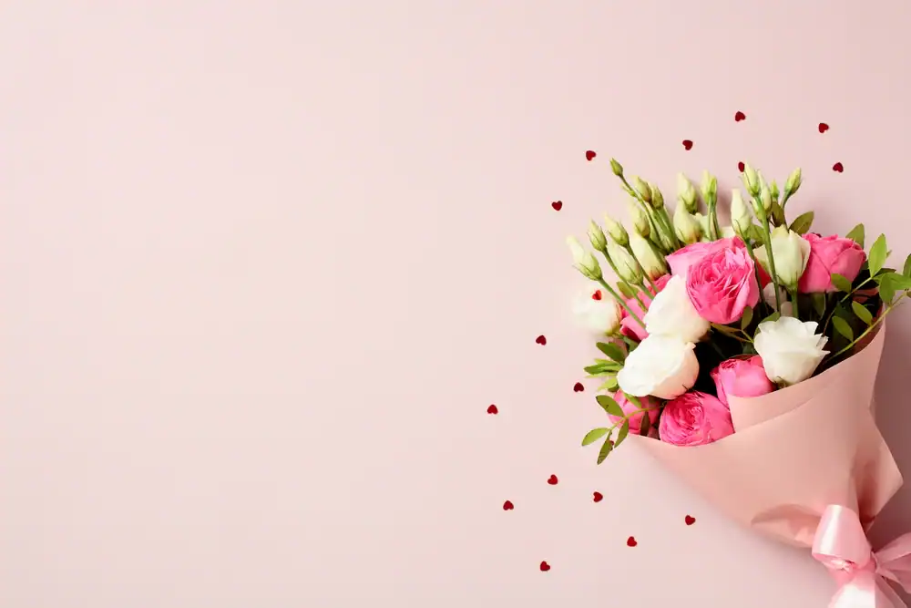 A bouquet of pink and white roses wrapped in pink paper is placed on a light pink background, with small red heart confetti scattered nearby.