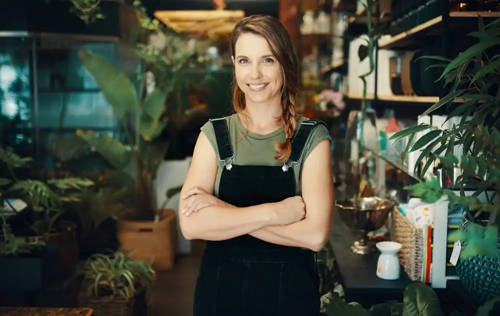 A woman with braided hair stands smiling with arms crossed in a plant-filled shop, wearing a green shirt and black overalls. The background shows shelves and a variety of potted plants.