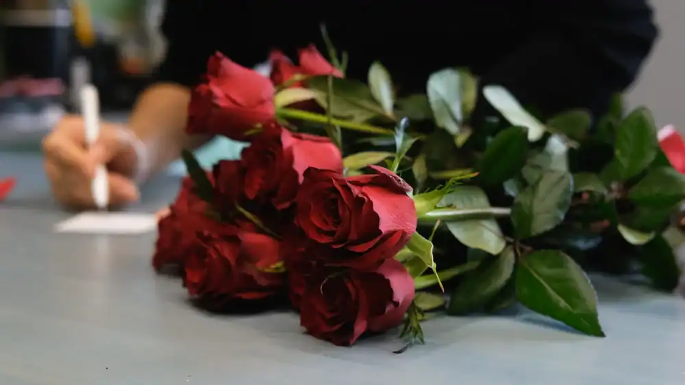 A bouquet of red roses rests on a table, while a person in the background writes on a piece of paper with a pen.