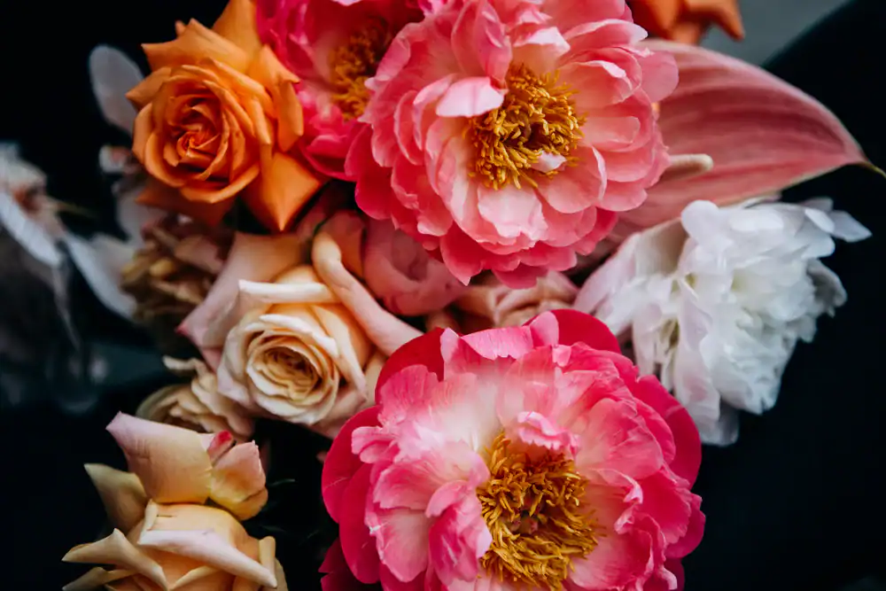 A close-up of a vibrant flower bouquet featuring large pink peonies, orange roses, white blooms, and green foliage against a dark background.