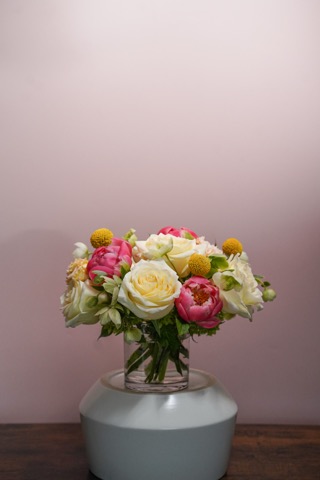 A glass vase with white, pink, and yellow flowers, including roses and billy balls, sits on a white round stand against a pale pink background on a wooden surface.
