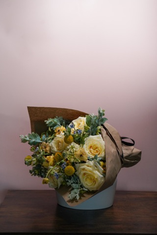 A bouquet of yellow and white flowers, including roses, wrapped in brown paper, sits on a wooden surface against a light pink background.