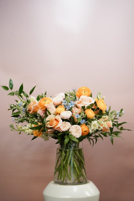 A clear glass vase filled with an arrangement of orange and pale pink roses, green leaves, and light blue flowers sits on a white surface against a soft pink background.