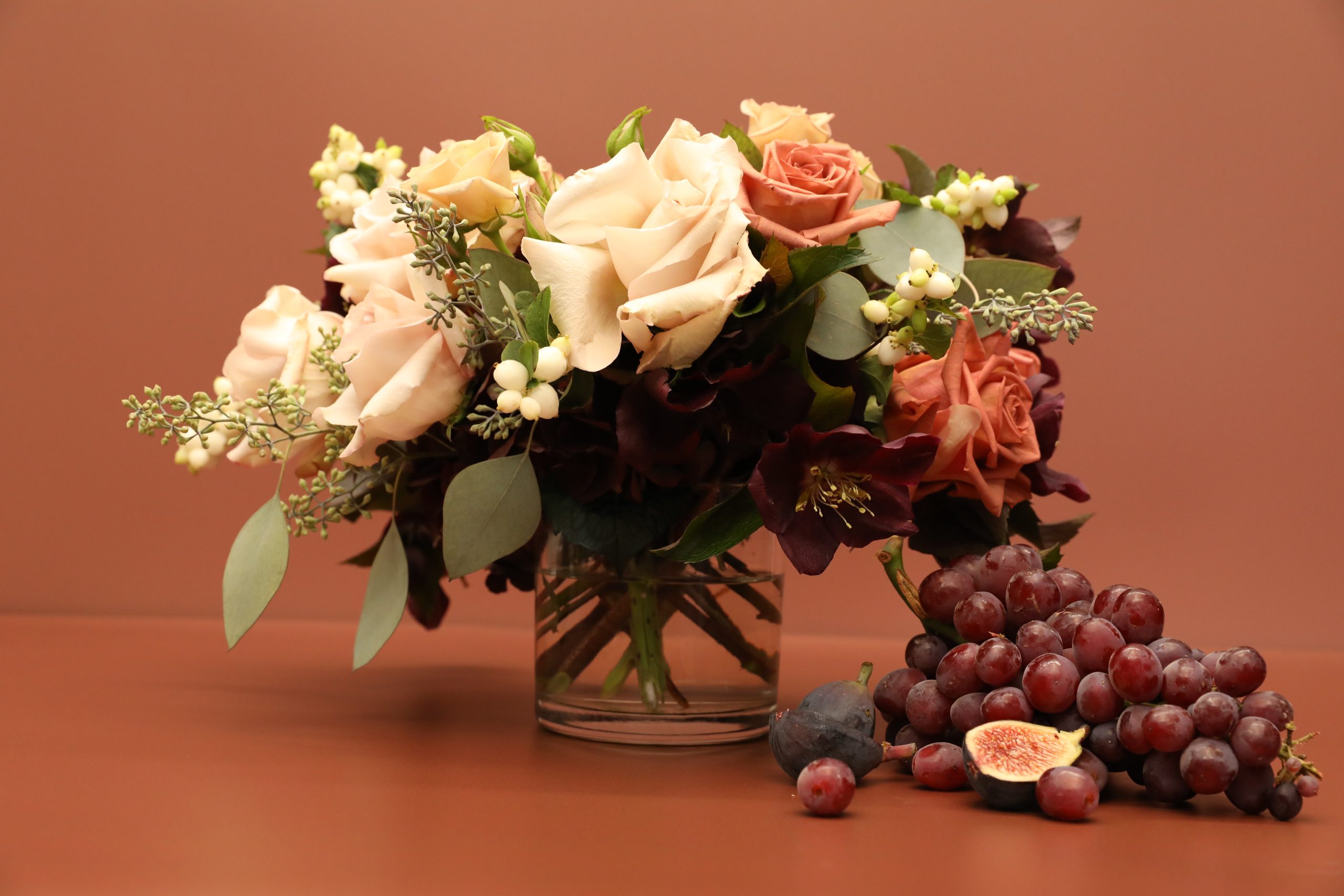 A glass vase filled with cream, blush, and dark red roses, green leaves, and white berries sits next to a cluster of red grapes and sliced figs on a brown surface with a matching brown background.