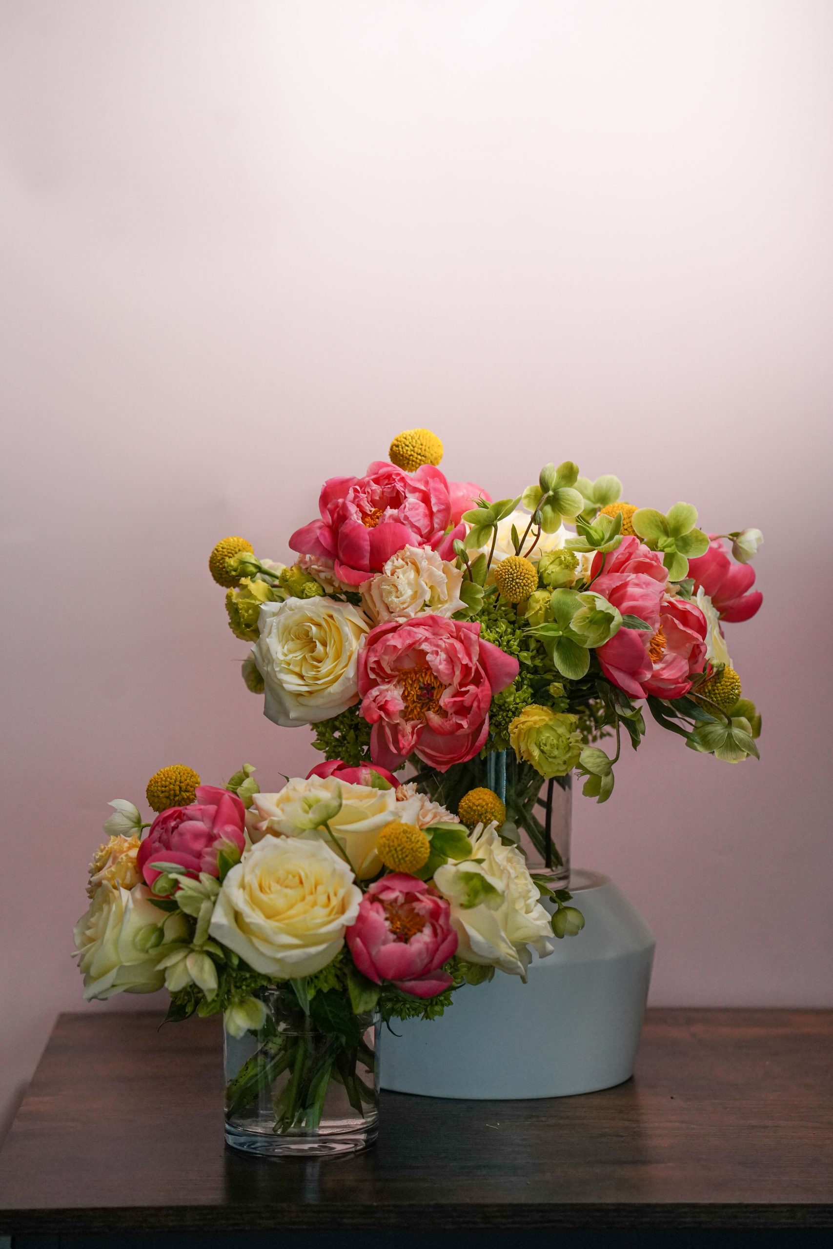 Three vases with vibrant flower arrangements, featuring pink peonies, yellow billy balls, white roses, and green hydrangeas, sit on a wooden table against a soft pink background.