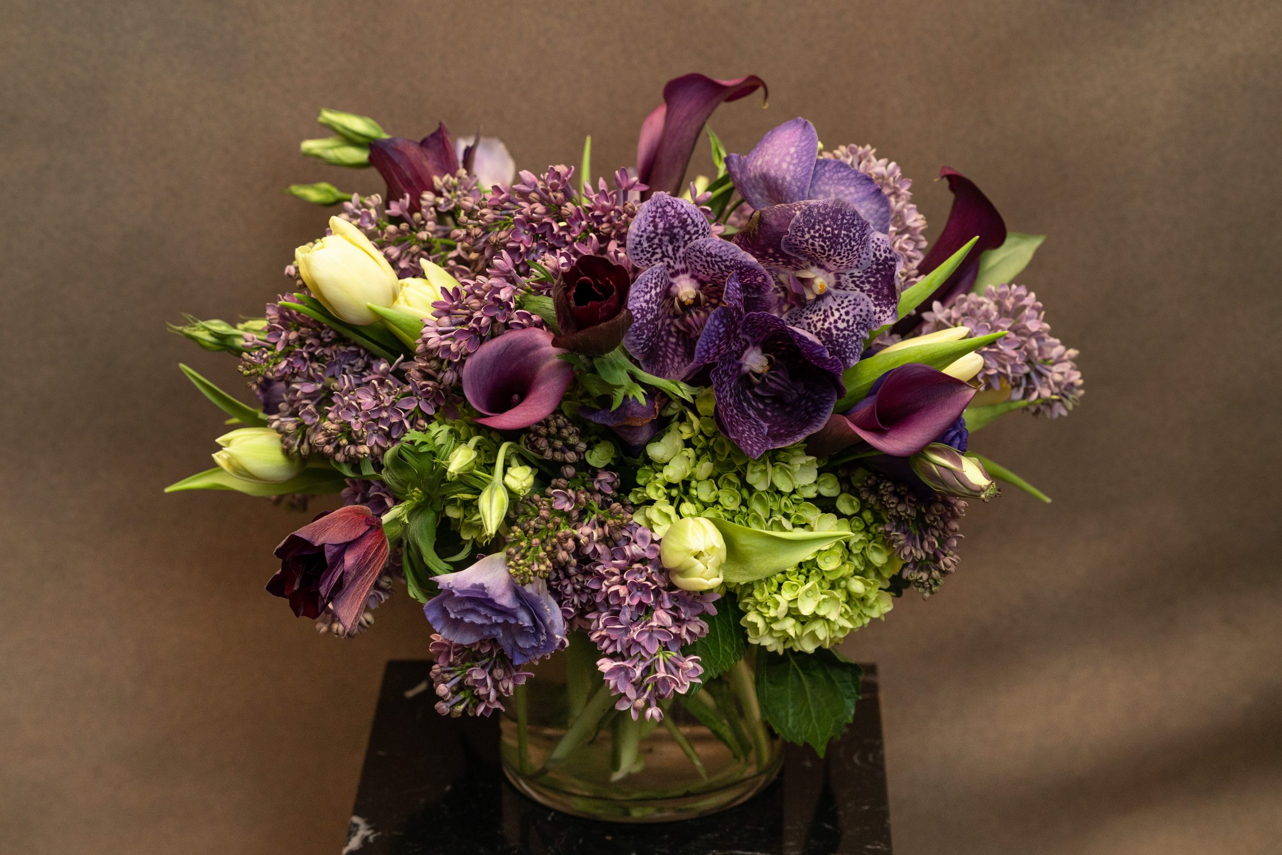 A floral arrangement in a glass vase featuring purple orchids, dark purple calla lilies, lilacs, pale green hydrangeas, white tulips, and assorted greenery against a brown background.